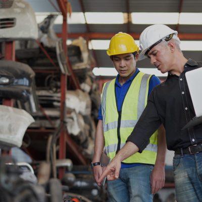 senior engineer using laptop computer checking list old used car engine with young asian worker in machinery parts shop warehouse. old manager find vehicle automobile machine in Recycle motor factory